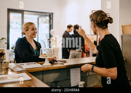 Donna anziana che parla con il cliente sorridente mentre indica uomo in caffetteria Foto Stock