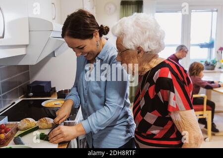 Vista laterale della donna anziana che guarda la figlia che taglia le zucchine in cucina Foto Stock