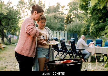 Vista laterale di madre e figlia che preparano cibo sopra barbecue griglia in cortile Foto Stock