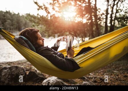 Contemplando libro di lettura dell'uomo mentre sdraiato sopra amaca dentro foresta Foto Stock