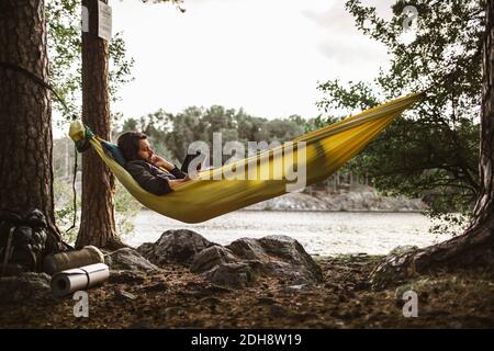 Libro di lettura del giovane uomo mentre sdraiato sopra amaca dentro foresta Foto Stock