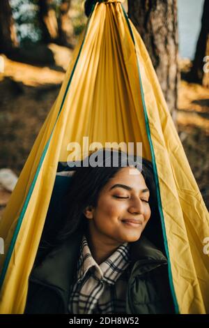 Donna sorridente con occhi chiusi su amaca in foresta durante vacanza Foto Stock