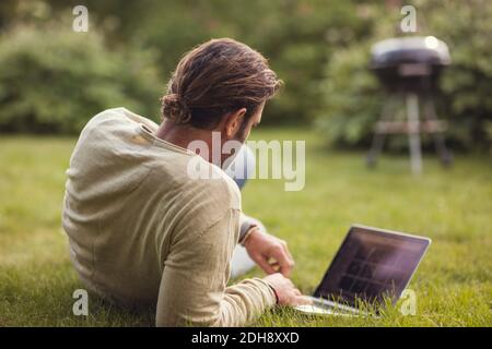 Vista posteriore dell'uomo reclinato con il computer portatile sull'erba cortile posteriore Foto Stock