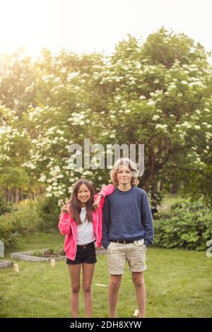 Ritratto di ragazza felice che mostra il segno di pace mentre si sta accanto amico nel cortile posteriore Foto Stock