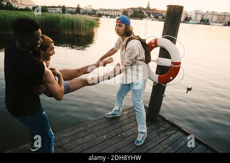 Amici maschili che gettano l'uomo nel lago mentre si sta sul molo durante il tramonto Foto Stock