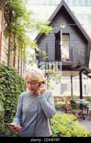 Donna d'affari matura che utilizza cuffie intrauricolari e smartphone in ufficio iarda Foto Stock