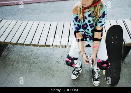 Vista ad alto angolo della ragazza con skateboard che tiene il telefono cellulare mentre si siede sulla panchina al parco Foto Stock