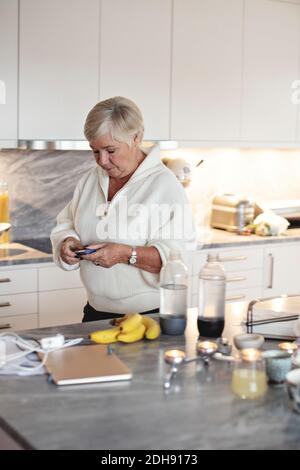 Donna anziana che tiene la scatola della pillola mentre si sta in piedi da isola della cucina Foto Stock