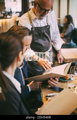 Cameriere in piedi da tavolo mentre i professionisti di affari si siedono nel ristorante Foto Stock