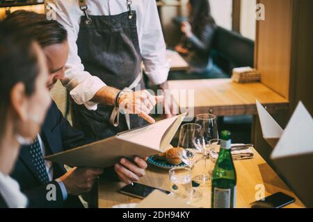 Sezione centrale del cameriere che indica il menu mentre i professionisti di affari si siedono nel ristorante Foto Stock