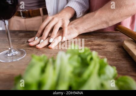 Vista ritagliata della donna che tocca la mano del ragazzo vicino al vetro di vino e lattuga in primo piano sfocato Foto Stock