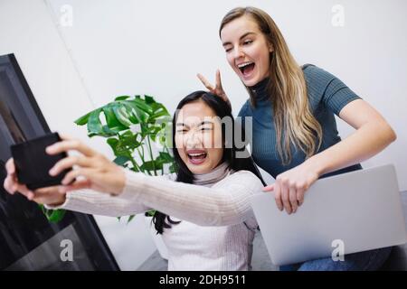 Allegro blogger femminile prendendo selfie attraverso smartphone in creativo ufficio Foto Stock