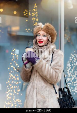 una donna in abiti invernali che guarda alla finestra, shopping e tenere una tazza di caffè Foto Stock