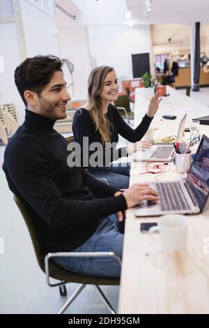 Giovane donna allegra e uomo che guarda via mentre si siede scrivania in ufficio Foto Stock