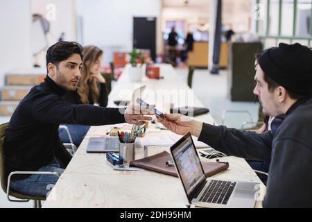Vista laterale di un giovane uomo che offre uno smartphone al collega alla scrivania in ufficio Foto Stock