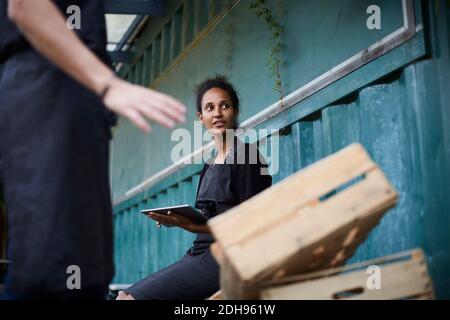 Donna giardiniere che tiene tablet digitale mentre amico gesturing da casse Foto Stock