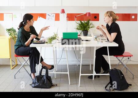 Tutta la lunghezza delle donne d'affari che parlano mentre pranzano al tavolo in ufficio Foto Stock