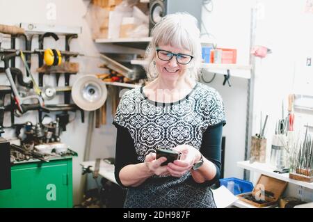 Donna anziana sorridente che usa il telefono cellulare in un'officina di gioielleria Foto Stock