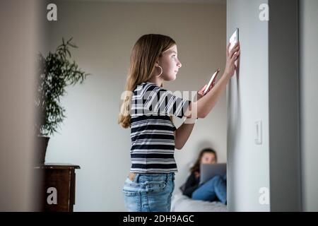 Ragazza che tiene il telefono cellulare mentre usa il tablet digitale sulla parete con sorella seduta in background in casa intelligente Foto Stock