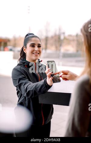 Cliente che accede allo smartphone durante la ricezione del pacchetto dalla consegna donna Foto Stock