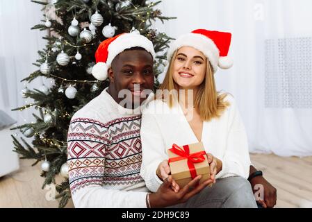 La coppia innamorata è seduta nel soggiorno festivo decorato di Natale e tenuta del regalo. Entrambi stanno guardando direttamente nella macchina fotografica felicemente. Allegra Christma Foto Stock