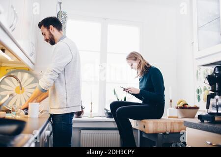 Vista laterale di piatti di lavaggio uomo mentre donna usa mobile telefono in cucina a casa Foto Stock