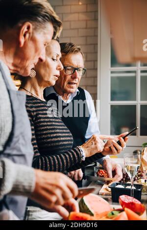 Donna anziana che discute con amici di sesso maschile su smartphone mentre in piedi in cucina a casa Foto Stock