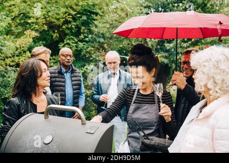 Sorridente femmina amici che cucinano la cena sulla griglia del barbecue durante la festa mentre piove Foto Stock