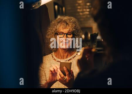 Donna anziana sorridente che usa lo smartphone mentre si siede a illuminato a casa durante la festa Foto Stock