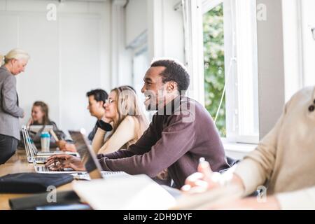 Vista laterale di un giovane sorridente utilizzando un computer portatile alla scrivania in classe Foto Stock