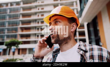 Uomo caucasico lavoratrice di costruzione in cappello arancione e camicia plaid che parla al telefono al cantiere. Tema dell'architettura. Foto Stock