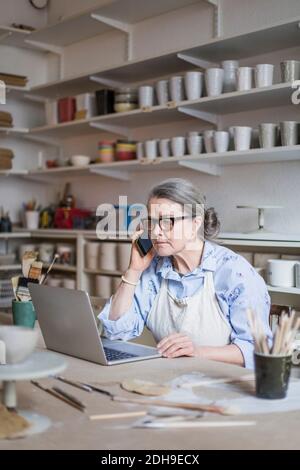 Femmina senior potter che parla sul telefono cellulare mentre si utilizza il computer portatile al tavolo in officina Foto Stock