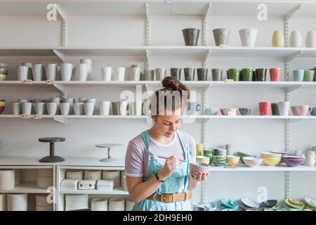 Giovane femmina vasaio usando l'attrezzo a mano sul vaso mentre si è in piedi contro gli scaffali del negozio Foto Stock