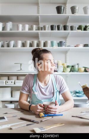 Giovane femmina vasaio seduto al banco da lavoro mentre si guarda lontano contro scaffali in negozio Foto Stock