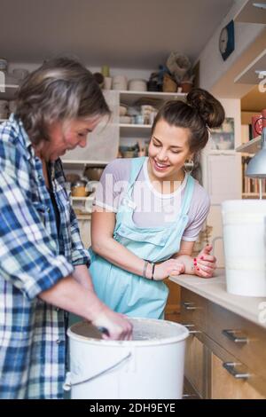 Donna felice che guarda il vasaio femmina maturo che mescola creta dentro benna in magazzino Foto Stock