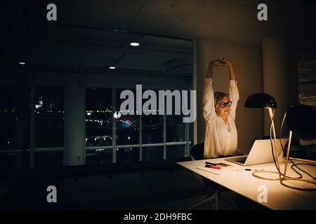 Donna d'affari esausta che allunga con le braccia sollevate mentre lavora tardi ufficio di lavoro illuminato Foto Stock