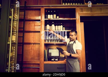 Proprietario maschile che controlla l'elenco delle bottiglie di vino mentre si è in piedi caffè Foto Stock