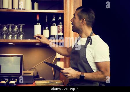 Uomo proprietario con tablet digitale guardando bottiglie di vino mentre in piedi nel cafe Foto Stock