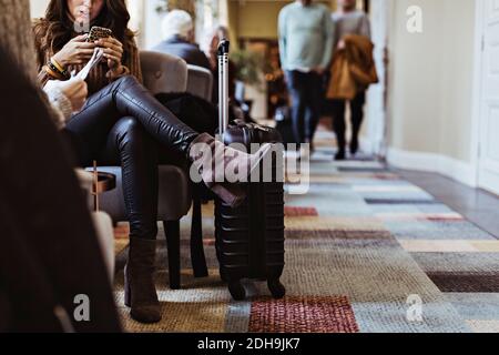 Sezione bassa della donna dell'adulto medio che usa il telefono mobile mentre sedersi sulla sedia nella hall dell'hotel Foto Stock