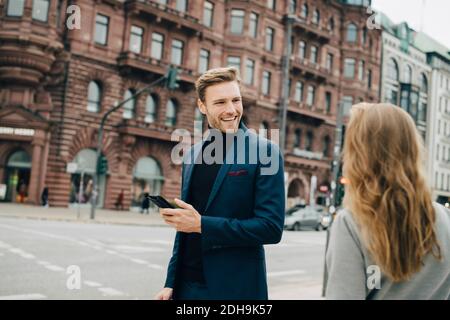 Uomo d'affari sorridente con il telefono che guarda la collega femmina mentre si è in piedi in città Foto Stock