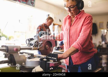 Donna anziana che usa la sega circolare mentre l'uomo sta in piedi sullo sfondo in officina Foto Stock