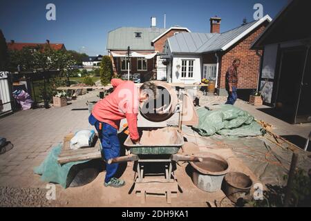 Donna anziana che usa il miscelatore del cemento al cantiere mentre l'uomo cammina in background Foto Stock