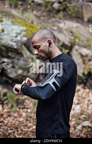 Vista laterale dell'atleta maschile che controlla l'orologio intelligente nella foresta Foto Stock