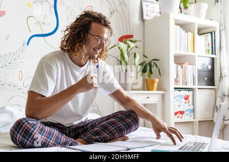 Foto di sorridente giovane bell'uomo al chiuso a casa scrittura note in notebook e studio online da computer portatile Foto Stock