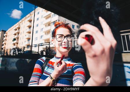 Giovane donna che guarda il telefono cellulare mentre si applica il rossetto città Foto Stock