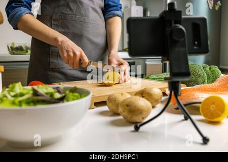 video di registrazione del blogger di cibo con la macchina fotografica del telefono alla cucina domestica. tagliando la patata sul bordo di legno Foto Stock