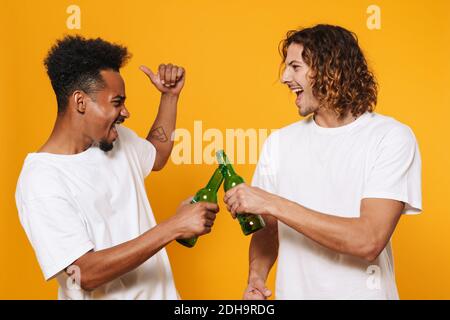 Eccitato multiculturale due ragazzi gridando e clinking bottiglie isolato sopra sfondo giallo Foto Stock