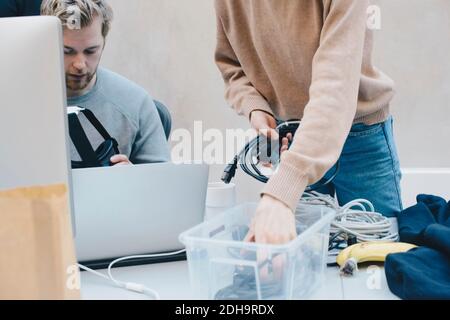 Sezione centrale del programmatore di computer femmina che rimuove i cavi dal contenitore mentre collega che tiene le cuffie in ufficio Foto Stock