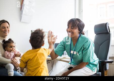 Un pediatra sorridente che dà cinque anni a ragazzo mentre la madre guarda affettuosamente in clinica Foto Stock