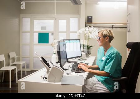 Vista laterale del medico femminile maturo che lavora sul computer mentre seduto in clinica Foto Stock